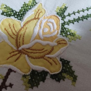May include: Close-up of white fabric featuring embroidered floral designs. A large yellow rose with white and brown detailing is the focal point. Green and blue floral embroidery is also visible, creating a handcrafted, vintage aesthetic.