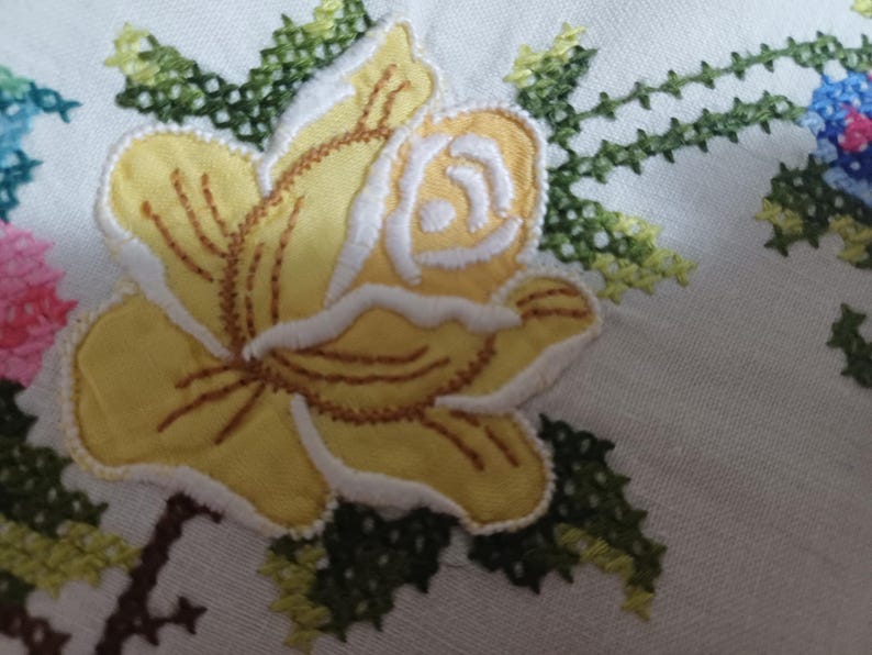 May include: Close-up of white fabric featuring embroidered floral designs. A large yellow rose with white and brown detailing is the focal point. Green and blue floral embroidery is also visible, creating a handcrafted, vintage aesthetic.