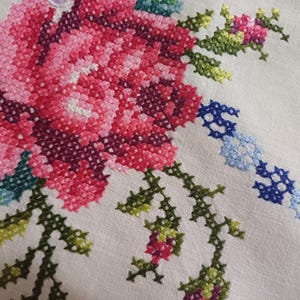 May include: Close-up of a cross-stitched floral design. The embroidery features a large pink and red rose with green leaves and stems. A blue and white chain-like pattern is also visible on the off-white fabric. The needlework is detailed and colourful.