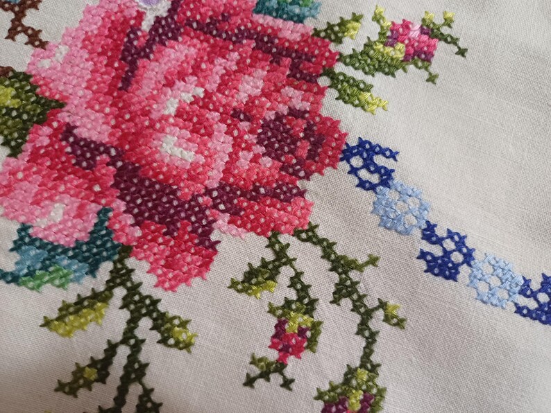 May include: Close-up of a cross-stitched floral design. The embroidery features a large pink and red rose with green leaves and stems. A blue and white chain-like pattern is also visible on the off-white fabric. The needlework is detailed and colourful.