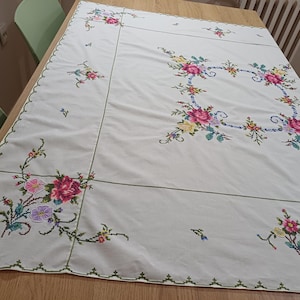 May include: White tablecloth with embroidered floral designs in pink, yellow, and purple. The embroidery features a square floral border and smaller floral clusters along the edges. The tablecloth is on a wooden table.