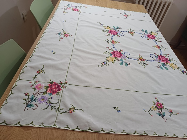 May include: White tablecloth with embroidered floral designs in pink, yellow, and purple. The embroidery features a square floral border and smaller floral clusters along the edges. The tablecloth is on a wooden table.