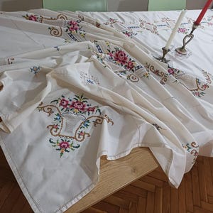 May include: A white tablecloth with intricate floral embroidery in pink, green, and blue. The design includes a basket of flowers and decorative borders. Two candle holders with candles are on the table.