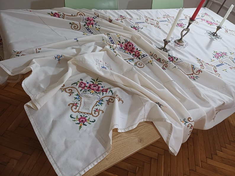 May include: A white tablecloth with intricate floral embroidery in pink, green, and blue. The design includes a basket of flowers and decorative borders. Two candle holders with candles are on the table.