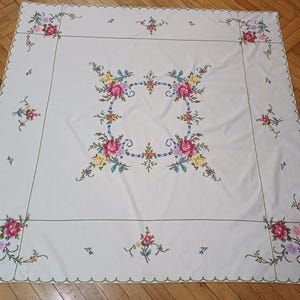 May include: A white tablecloth with colourful floral embroidery. The embroidery features pink, yellow, and purple roses, with green leaves and stems. The tablecloth has a scalloped edge and is laid out on a wooden floor.