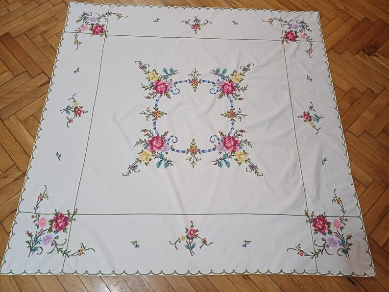 May include: A white tablecloth with colourful floral embroidery. The embroidery features pink, yellow, and purple roses, with green leaves and stems. The tablecloth has a scalloped edge and is laid out on a wooden floor.