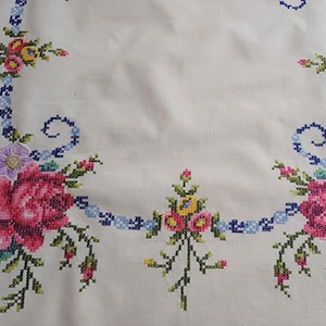 May include: White tablecloth with cross-stitched floral embroidery. The embroidery features pink roses, yellow and blue flowers, and green leaves. A blue ribbon-like design frames the floral arrangements.