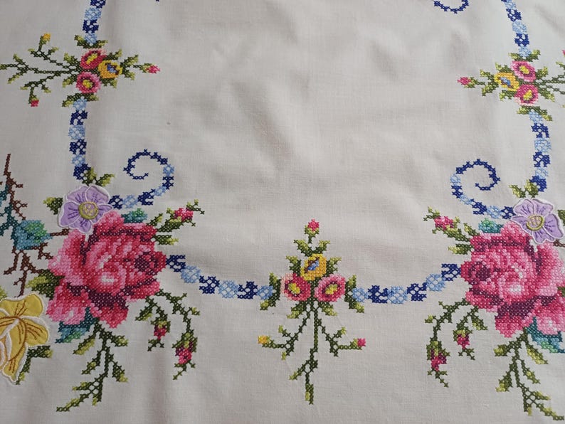 May include: White tablecloth with cross-stitched floral embroidery. The embroidery features pink roses, yellow and blue flowers, and green leaves. A blue ribbon-like design frames the floral arrangements.