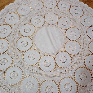 May include: A round, white lace tablecloth with a detailed, openwork design. The tablecloth features a central section and a border of repeating circular patterns, creating a delicate and elegant aesthetic. The intricate lace adds a vintage or handmade feel.
