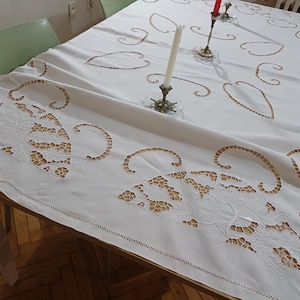 Pode incluir: Uma toalha de mesa retangular branca com desenhos recortados intrincados e costuras decorativas. A toalha de mesa apresenta um padrão repetido de formas semelhantes a folhas e é drapeada sobre uma mesa de madeira. Dois castiçais com velas vermelhas e brancas estão sobre a mesa.