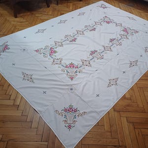 May include: A white tablecloth with intricate floral embroidery in pink, red, and green. The rectangular cloth features a central design and repeating patterns, creating a vintage aesthetic. The tablecloth is laid out on a wooden floor.
