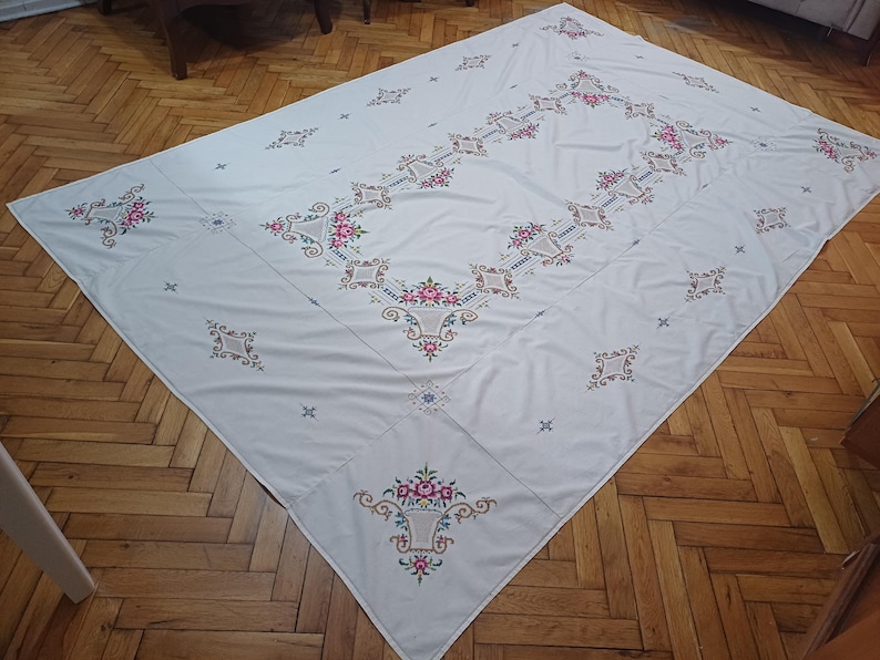 May include: A white tablecloth with intricate floral embroidery in pink, red, and green. The rectangular cloth features a central design and repeating patterns, creating a vintage aesthetic. The tablecloth is laid out on a wooden floor.