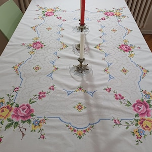 May include: White tablecloth with embroidered floral design. The embroidery features pink, yellow, and blue flowers with green stems. A decorative border and a central design element are also present. Two candlesticks with red and white candles are on the table.