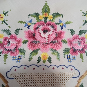 May include: Close-up of a white tablecloth with a floral cross-stitch design. The design features pink roses, green leaves, and accents of yellow and blue. The tablecloth has a scalloped edge with a lace insert.