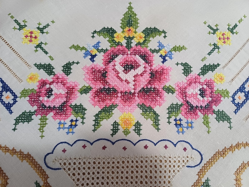 May include: Close-up of a white tablecloth with a floral cross-stitch design. The design features pink roses, green leaves, and accents of yellow and blue. The tablecloth has a scalloped edge with a lace insert.