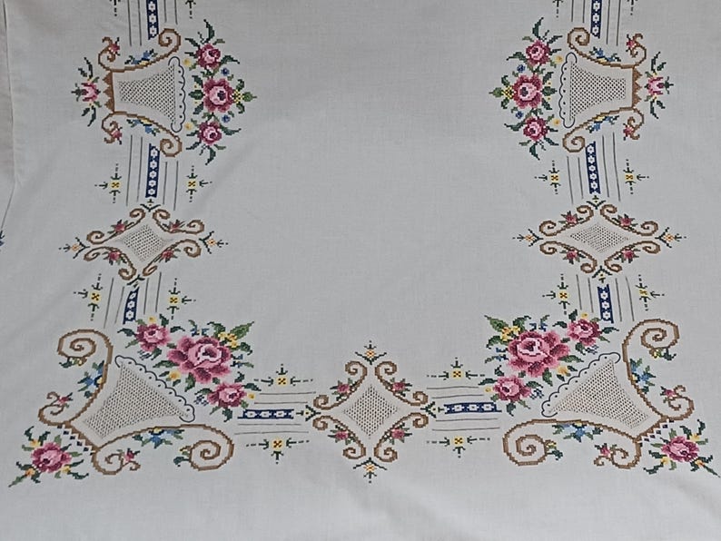 May include: White tablecloth with embroidered floral and basket designs. The embroidery features pink roses, green leaves, and accents of blue, brown, and yellow. The design is symmetrical, with floral arrangements in each corner and along the edges.