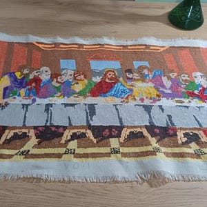 Op de afbeelding: Een tapijt dat het Laatste Avondmaal uitbeeldt, met Jezus en zijn discipelen aan een tafel. Het kunstwerk gebruikt verschillende kleuren, waaronder blauw, rood, geel en bruin. Het tapijt heeft een gefranjerde rand en ligt op een houten oppervlak.