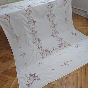 May include: White tablecloth with intricate floral embroidery in pink, brown, and blue. The embroidered design features repeating floral patterns and geometric shapes, creating a decorative and elegant look. The tablecloth is draped over a table.