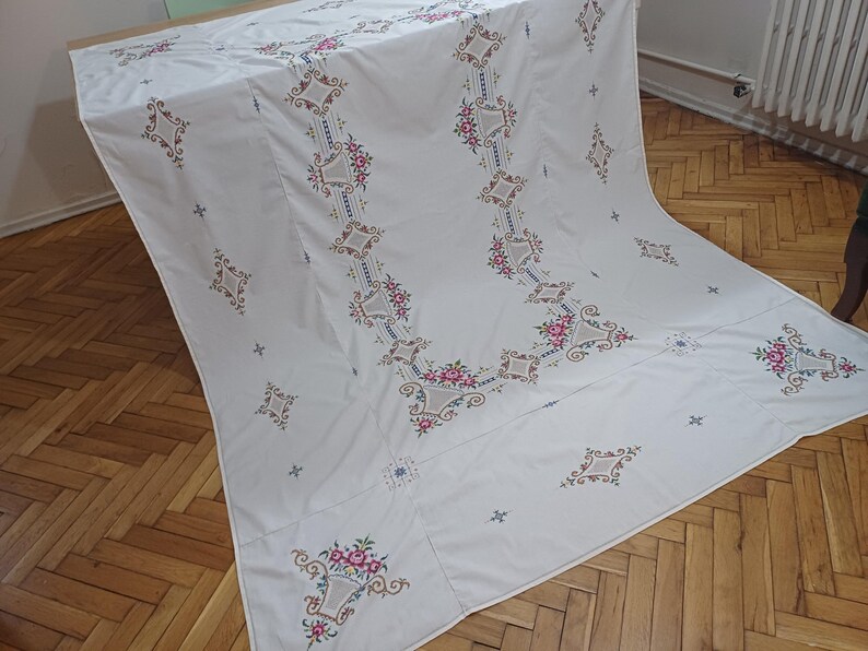 May include: White tablecloth with intricate floral embroidery in pink, brown, and blue. The embroidered design features repeating floral patterns and geometric shapes, creating a decorative and elegant look. The tablecloth is draped over a table.