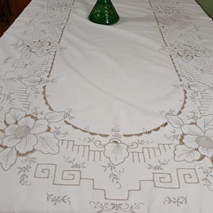 Può includere: Tovaglia bianca con ricami floreali e geometrici. Il ricamo presenta grandi fiori, foglie e motivi intricati in un colore neutro. Un vaso di vetro verde è sul tavolo. Adatta per cene o occasioni speciali.