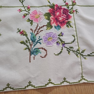 May include: White linen tablecloth with cross-stitched floral embroidery. The design features pink, purple, and red flowers with green stems and leaves. The border is trimmed with a green cross-stitch pattern.