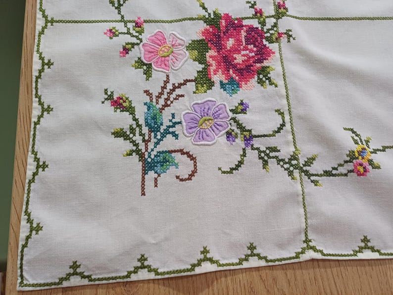 May include: White linen tablecloth with cross-stitched floral embroidery. The design features pink, purple, and red flowers with green stems and leaves. The border is trimmed with a green cross-stitch pattern.