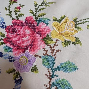 May include: Close-up of white fabric with colourful cross-stitch embroidery. The design features a large pink rose, a yellow rose, and a purple flower, with green leaves and blue accents. The embroidery is detailed and vibrant.
