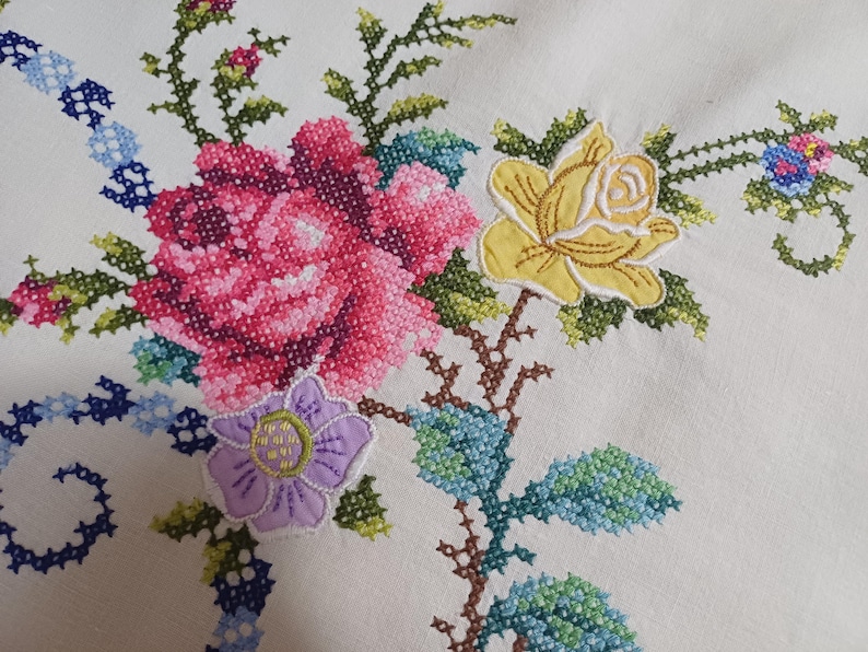 May include: Close-up of white fabric with colourful cross-stitch embroidery. The design features a large pink rose, a yellow rose, and a purple flower, with green leaves and blue accents. The embroidery is detailed and vibrant.