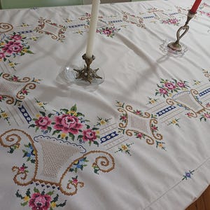 May include: White tablecloth with embroidered floral and basket designs. The embroidery features pink roses, blue and yellow accents, and brown basket details. The tablecloth is on a table with candle holders.