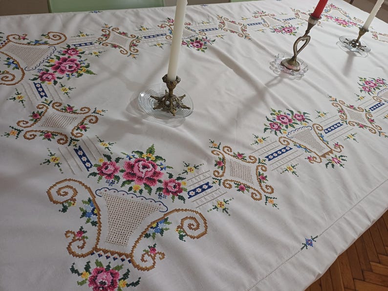 May include: White tablecloth with embroidered floral and basket designs. The embroidery features pink roses, blue and yellow accents, and brown basket details. The tablecloth is on a table with candle holders.