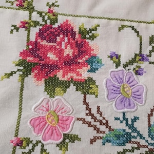 May include: Close-up of white fabric with colourful cross-stitch embroidery. The design features a large pink and purple flower, smaller pink and purple flowers, and green leaves. The embroidery is detailed and vibrant.