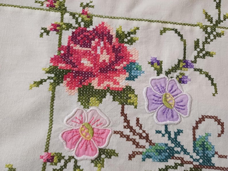 May include: Close-up of white fabric with colourful cross-stitch embroidery. The design features a large pink and purple flower, smaller pink and purple flowers, and green leaves. The embroidery is detailed and vibrant.