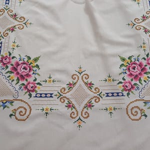 May include: White tablecloth with embroidered floral and geometric patterns. The design features pink roses, blue and yellow accents, and brown scrollwork. The embroidery is detailed and covers the edges of the cloth.