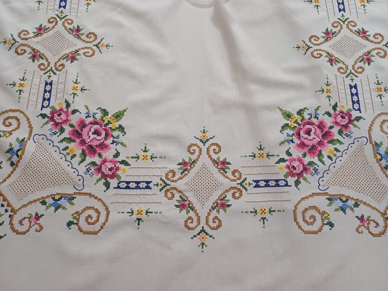 May include: White tablecloth with embroidered floral and geometric patterns. The design features pink roses, blue and yellow accents, and brown scrollwork. The embroidery is detailed and covers the edges of the cloth.