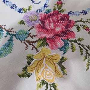 May include: Close-up of white fabric with colourful cross-stitch floral embroidery. The design features a large pink rose, a yellow flower, and a purple flower, with green and blue leaves and stems. The embroidery is detailed and vibrant.