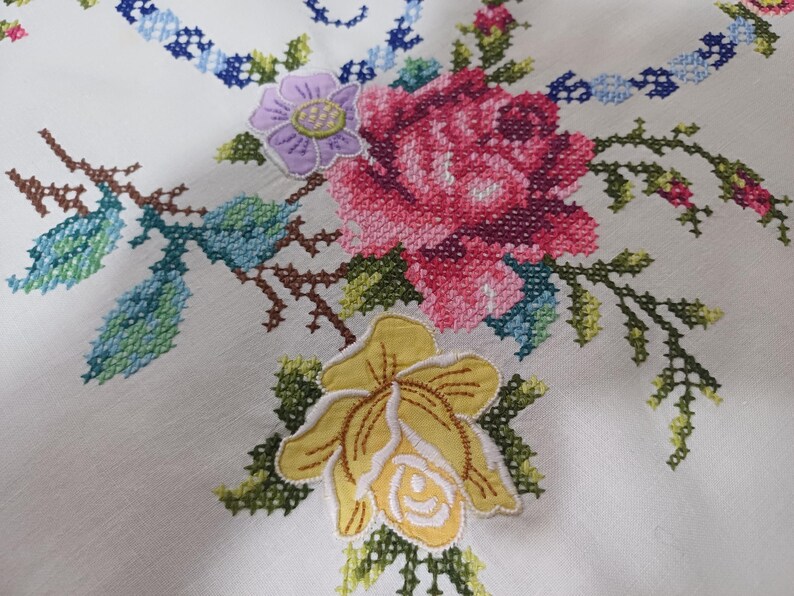 May include: Close-up of white fabric with colourful cross-stitch floral embroidery. The design features a large pink rose, a yellow flower, and a purple flower, with green and blue leaves and stems. The embroidery is detailed and vibrant.