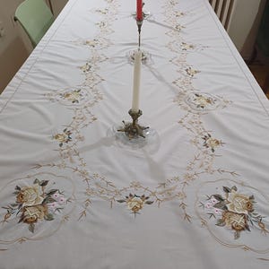 May include: A white tablecloth with embroidered floral designs in shades of beige, brown, and pink. The tablecloth is laid out on a table, with two candles and a candlestick in the center. The overall theme is home decor.