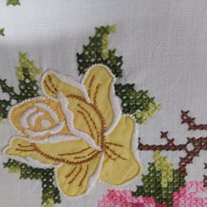 May include: Close-up of white fabric featuring embroidered floral designs. A large yellow rose with white edging is central, surrounded by green leaves and stems. Other flowers are pink, blue, and green.
