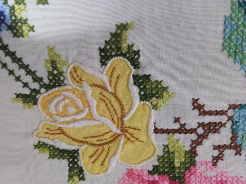 May include: Close-up of white fabric featuring embroidered floral designs. A large yellow rose with white edging is central, surrounded by green leaves and stems. Other flowers are pink, blue, and green.