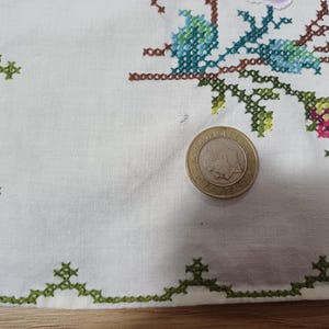 May include: Cream-coloured linen cloth with a green cross-stitch border and floral embroidery in blue, pink, and green. A coin is on the cloth. The embroidery features a detailed floral design.