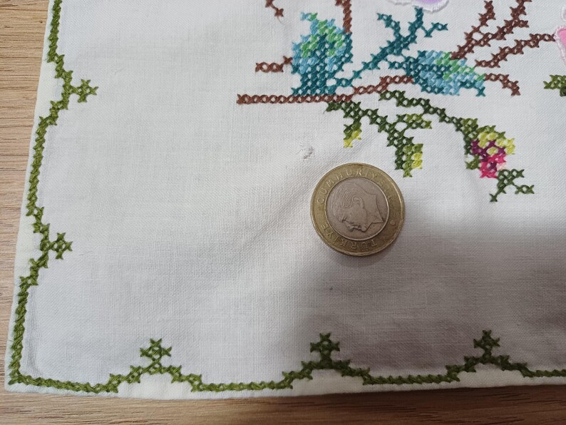 May include: Cream-coloured linen cloth with a green cross-stitch border and floral embroidery in blue, pink, and green. A coin is on the cloth. The embroidery features a detailed floral design.