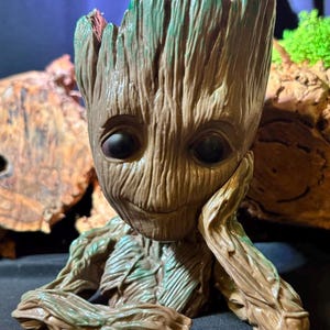 May include: A detailed figurine of Groot, a character from Guardians of the Galaxy. The figure is primarily brown with green accents, featuring a textured, tree-like appearance. Groot is depicted with a smiling expression, resting his chin on his hand.