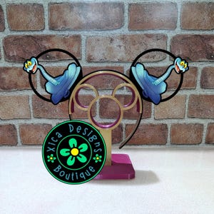 May include: A decorative headband stand featuring a Mickey Mouse ear design in gold and purple. The stand holds a pair of black-rimmed ears with a cartoon snake design. The stand has a purple base and a green and black logo that says "Xica Designs Boutique."
