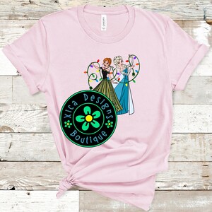 Peut inclure: T-shirt rose clair avec un dessin anim&eacute; de deux personnages tenant des guirlandes lumineuses. Les personnages portent des robes noires, vertes et bleues. Le t-shirt porte &eacute;galement un logo circulaire avec le texte "Xica Designs Boutique".