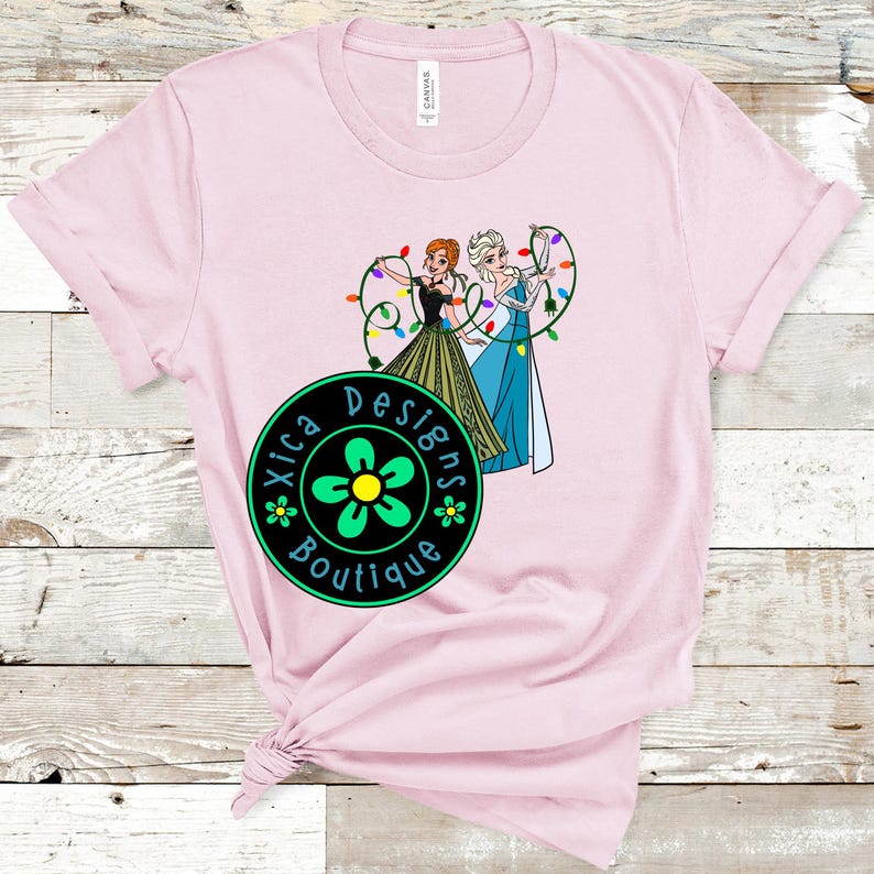 Peut inclure: T-shirt rose clair avec un dessin anim&eacute; de deux personnages tenant des guirlandes lumineuses. Les personnages portent des robes noires, vertes et bleues. Le t-shirt porte &eacute;galement un logo circulaire avec le texte "Xica Designs Boutique".