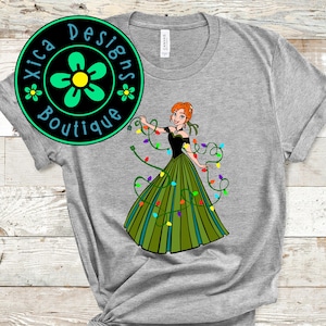 Pu&ograve; includere: Una maglietta grigia con un'immagine di cartone animato verde, nero e rosso di una donna che indossa un vestito verde con luci di Natale avvolte intorno a lei. Il testo "Xica Designs Boutique" &egrave; in un cerchio verde con un fiore giallo.