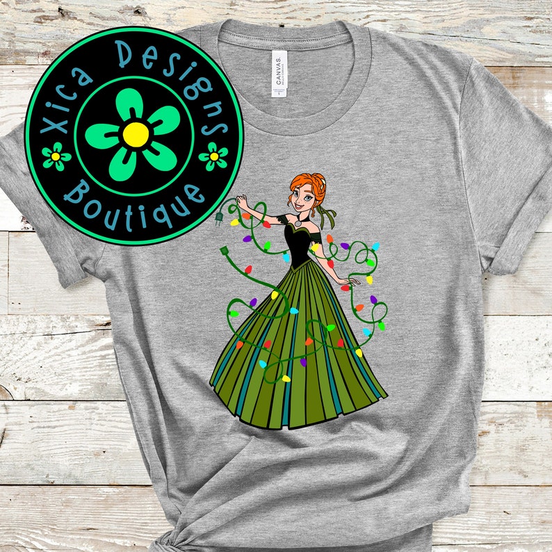 Pu&ograve; includere: Una maglietta grigia con un'immagine di cartone animato verde, nero e rosso di una donna che indossa un vestito verde con luci di Natale avvolte intorno a lei. Il testo "Xica Designs Boutique" &egrave; in un cerchio verde con un fiore giallo.