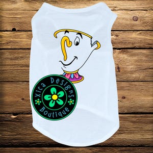 May include: White dog shirt featuring a cartoon teacup design with yellow, pink, and blue accents. The shirt includes a circular logo with the text "Xica Designs Boutique" and a green flower.