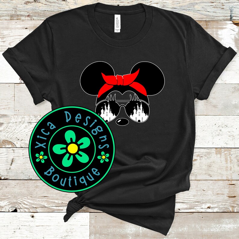 May include: Black t-shirt with a white silhouette of Mickey Mouse wearing sunglasses with a castle design. The sunglasses are reflected in the castle design. Mickey Mouse is wearing a red headband. The shirt has a green circle with a yellow flower and the text "Xica Designs Boutique" in white.