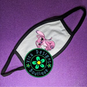 Puede incluir: Una mascarilla blanca con ribete y presillas negras. La mascarilla presenta un personaje de dibujos animados rosa. Un logotipo circular con el texto "Xica Designs Boutique" est&aacute; en la parte inferior.